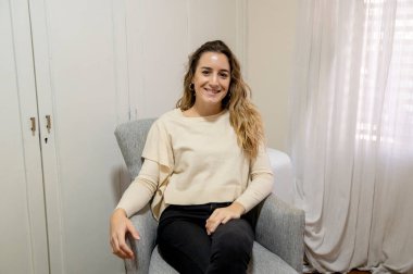 Evdeki kanepede oturan bir kadın kameraya bakıp gülümsüyor..