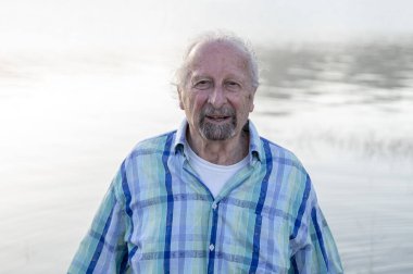 Adam kameraya bakıyor ve gülümsüyor. Doğada, gölün kenarında dikilirken..