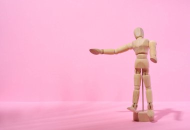 Wooden puppet man on a pink background, pointing with his hand