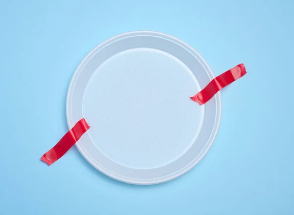 Empty white plastic plate attached with red adhesive tape on a blue background, a concept of refusing plastic tableware and recycling materials, zero waste