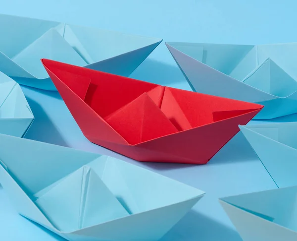 A red paper boat stands in front of a group of blue paper boats, a confrontation