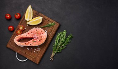 Tahta kesme tahtasında çiğ somon bifteği, limon dilimleri, baharatlar. Siyah masanın üstü. Boşluğu kopyala