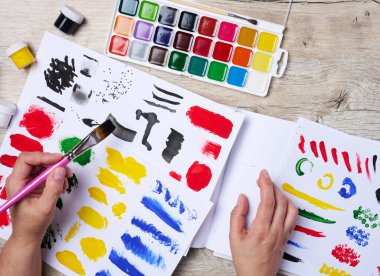 Bir dizi suluboya boya, fırça ve farklı renkte kağıt. Fırça tutan kadın eller