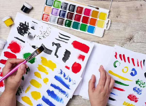 Bir dizi suluboya boya, fırça ve farklı renkte kağıt. Fırça tutan kadın eller
