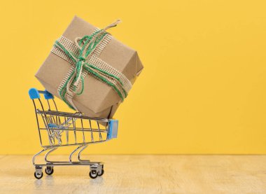 Kahverengi el sanatları kağıdına sarılı bir hediye. Minyatür bir alışveriş arabasında, sarı arka planda.