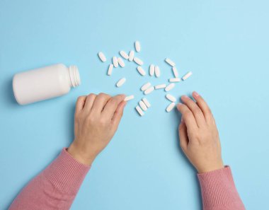 Mavi arka planda plastik bir şişe ve beyaz oval haplar tutan bir kadın eli..