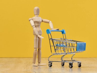 Ahşap bir manken sarı arka planda boş bir alışveriş arabası bulundurur. İnternetten alışveriş yapma ve ürün sipariş etme kavramını temsil eder.