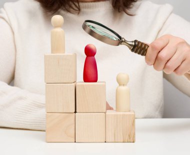 Woman holding a magnifying glass and wooden men on a white table. Personnel recruitment concept, talented employees. Career advancement
