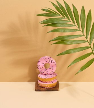 Pembe jöleli ve çok renkli serpiştirilmiş donut, palmiye yaprağından bir gölgeyle kahverengi arka plan