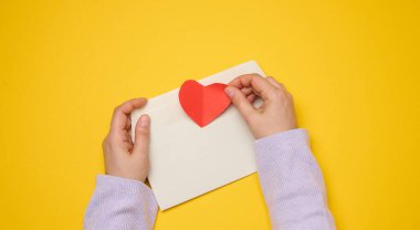 Dişi eller sarı arka planda beyaz dikdörtgen bir zarf tutar, kağıt kırmızı kalpler. Yukarıdan görüntüle