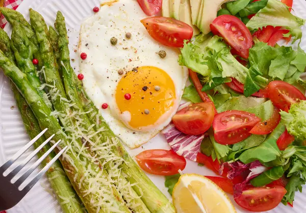 Yuvarlak tabak, pişmiş kuşkonmaz, sahanda yumurta, avokado ve taze sebze salatası.