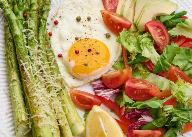 Yuvarlak tabak, pişmiş kuşkonmaz, sahanda yumurta, avokado ve taze sebze salatası.