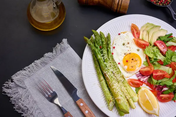 Yuvarlak tabak, pişmiş kuşkonmaz, sahanda yumurta, avokado ve taze sebze salatası.