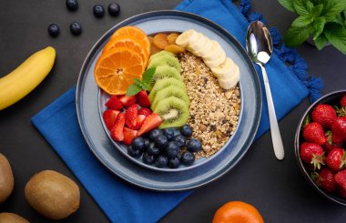 Çilekli granola, kivi, muz ve yaban mersini siyah bir masada yuvarlak bir tabakta. Sağlıklı ve lezzetli yemekler.