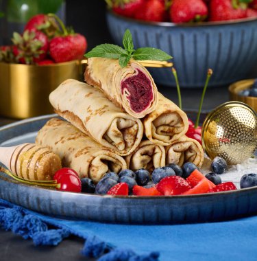 Yığınla krep yığılmış, bir tanesi kesilmiş ve böğürtlen dolgusu ile dolu. Tabak ayrıca böğürtlen, çilek ve kirazla süslenmiş, ballı bir ballı dipper, nane filizi ve altın elek ile süslenmiş. Koyu mavi bir masa örtüsü..