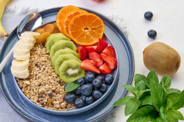 Beyaz bir masada yuvarlak bir tabakta çilek, kivi, muz ve yaban mersinli granola. Sağlıklı ve lezzetli yemekler.