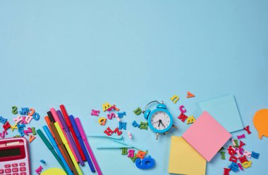 Kırtasiye malzemesi mavi arka planda. Çok renkli keçeli kalemler, hesap makinesi, alarm saati, yapışkan notlar ve ahşap harfler, çalışmaya ya da çalışmaya hazırlanma, çalışma alanını düzenleme