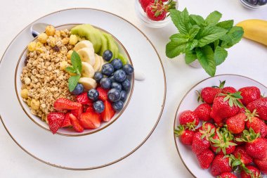 Beyaz bir masada yuvarlak bir tabakta çilek, kivi, muz ve yaban mersinli granola. Sağlıklı ve lezzetli yemekler.