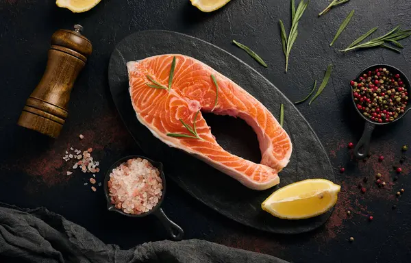 Oval siyah tabakta çiğ somon bifteği ve koyu arkaplanda limon dilimleri. Üst görünüm
