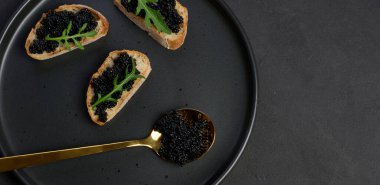 Üç parça çıtır ekmek, lüks siyah havyar ve taze roka, şık bir siyah tabakta servis ediliyor. Boşluğu kopyala