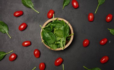 Açılmış kiraz domatesleri ve koyu arkaplanda ıspanak ile çevrili taze ıspanak yapraklarıyla dolu ahşap bir kase.