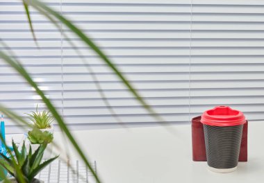 Arka planda beyaz perdeleri olan ofis masası. Gri ve kırmızı kahve fincanı, yeşil bitki ve tel örgüyle birlikte masanın üzerinde duruyor. Masa temiz ve minimalist..