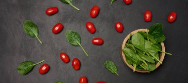 Açılmış kiraz domatesleri ve koyu arkaplanda ıspanak ile çevrili taze ıspanak yapraklarıyla dolu ahşap bir kase.