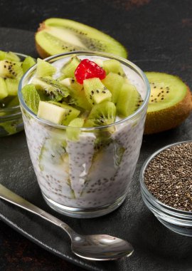 Bir bardak kivi dilimlenmiş chia tohumu pudingi ve kırmızı kivi koyu bir yüzeyde yarım kivi, bir kase doğranmış kivi ve chia tohumuyla duruyor.