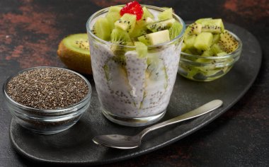 Bir bardak kivi dilimlenmiş chia tohumu pudingi ve kırmızı kivi koyu bir yüzeyde yarım kivi, bir kase doğranmış kivi ve chia tohumuyla duruyor.