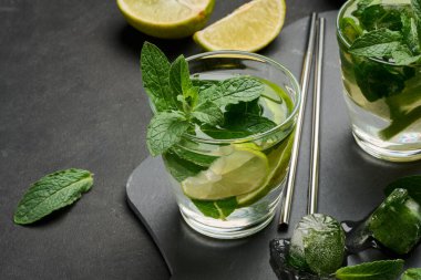 Bir bardak mojito kokteyli, taze nane yaprakları, limon dilimleri ve koyu taştan bir arka planda buz. Paslanmaz çelik pipetlerle ferahlatıcı yaz içeceği