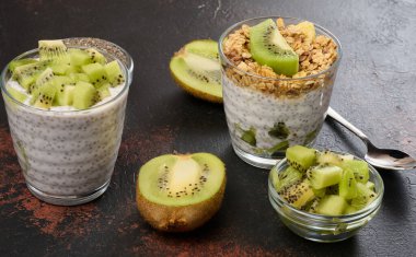İki bardak chia tohumu pudingi, biri doğranmış kivi, diğeri granola ve muz cipsi ile kaplı, koyu bir yüzeyde malzemelerle birlikte.
