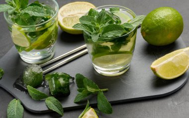 Bir bardak mojito kokteyli, taze nane yaprakları, limon dilimleri ve koyu taştan bir arka planda buz. Paslanmaz çelik pipetlerle ferahlatıcı yaz içeceği