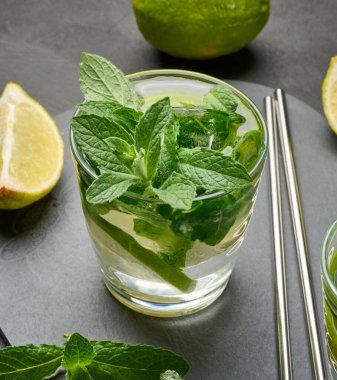 Bir bardak mojito kokteyli, taze nane yaprakları, limon dilimleri ve koyu taştan bir arka planda buz. Paslanmaz çelik pipetlerle ferahlatıcı yaz içeceği