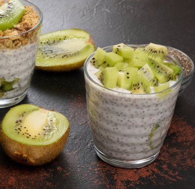 İki bardak chia tohumu pudingi, biri doğranmış kivi, diğeri granola ve muz cipsi ile kaplı, koyu bir yüzeyde malzemelerle birlikte.