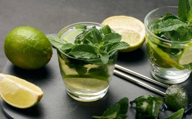 Bir bardak mojito kokteyli, taze nane yaprakları, limon dilimleri ve koyu taştan bir arka planda buz. Paslanmaz çelik pipetlerle ferahlatıcı yaz içeceği