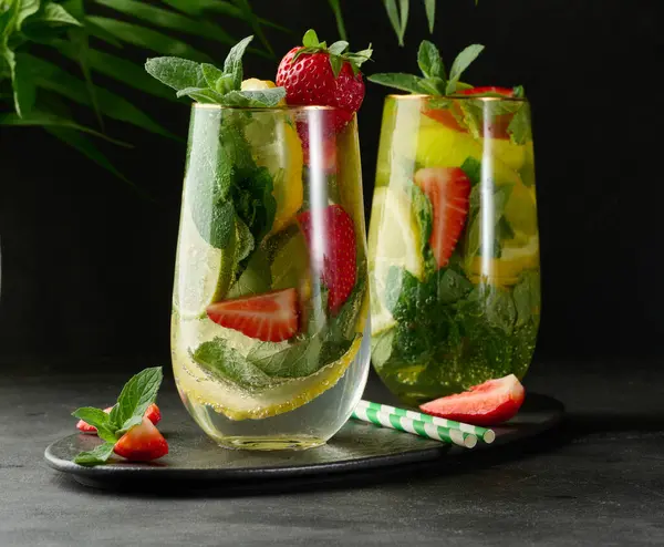 Limon dilimleri, çilekler, taze nane yaprakları ve siyah bir tabakta limon ve nane ile servis edilen köpüklü su dolu bir bardakta ferahlatıcı bir içecek.