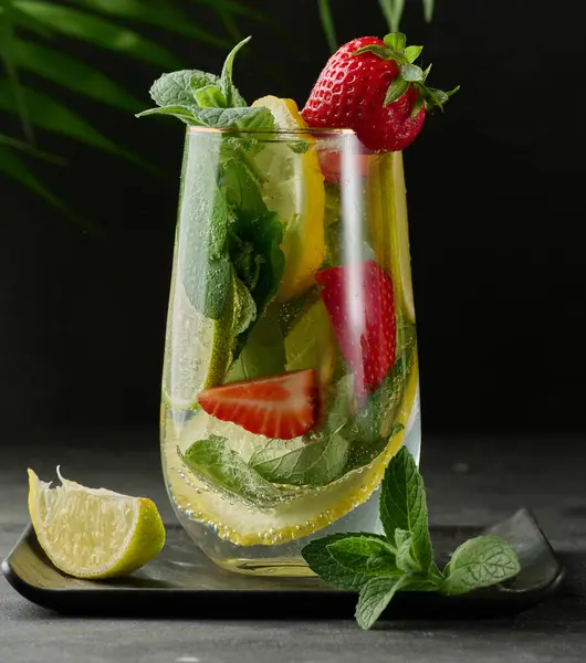 Limon dilimleri, çilekler, taze nane yaprakları ve siyah bir tabakta limon ve nane ile servis edilen köpüklü su dolu bir bardakta ferahlatıcı bir içecek..