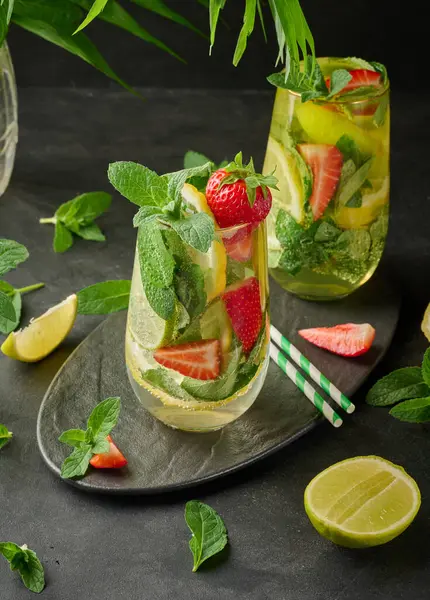 Limon dilimleri, çilekler, taze nane yaprakları ve siyah bir tabakta limon ve nane ile servis edilen köpüklü su dolu bir bardakta ferahlatıcı bir içecek.