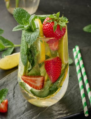 Limon dilimleri, çilekler, taze nane yaprakları ve siyah bir tabakta limon ve nane ile servis edilen köpüklü su dolu bir bardakta ferahlatıcı bir içecek.