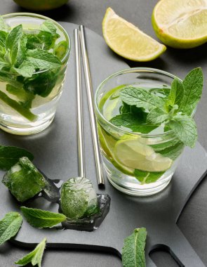 Bir bardak mojito kokteyli, taze nane yaprakları, limon dilimleri ve koyu taştan bir arka planda buz. Paslanmaz çelik pipetlerle ferahlatıcı yaz içeceği