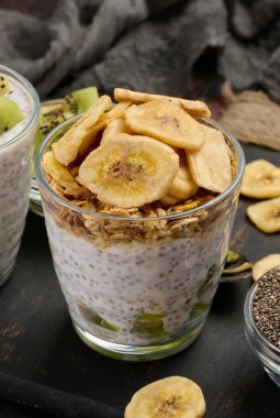İki bardak chia tohumu pudingi, biri doğranmış kivi, diğeri granola ve muz cipsi ile kaplı, koyu bir yüzeyde malzemelerle birlikte.