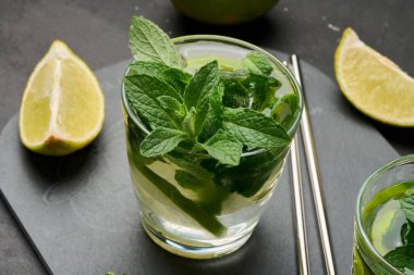 Bir bardak mojito kokteyli, taze nane yaprakları, limon dilimleri ve koyu taştan bir arka planda buz. Paslanmaz çelik pipetlerle ferahlatıcı yaz içeceği