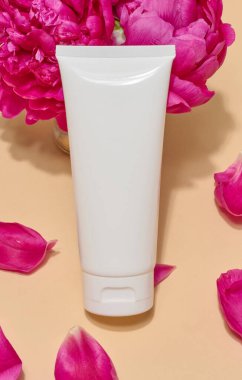 Üzerinde parlak pembe şakayıklar ve bej arka planda taç yaprakları olmayan beyaz kozmetik tüplerin yakın plan görüntüleri..