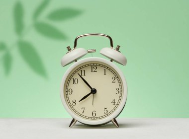 Beyaz antika çalar saat, yaprakların gölgeleriyle yeşil bir zeminde duruyor. Uyanıyorum ve güne başlıyorum.