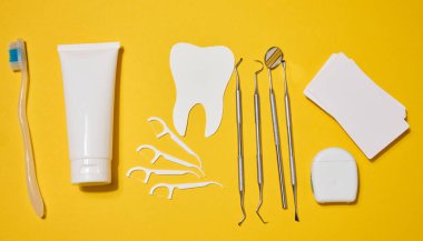 Beyaz diş macunu tüpü, diş fırçası, diş aletleri, plastik diş ipi, ve parlak sarı arka planda kesilmiş diş ipi. Üst görünüm