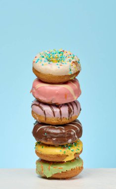 Değişik malzemelerle dolu altı renkli, jöleli donuttan oluşan uzun bir yığın, açık mavi arka planda beyaz bir yüzeye merkezi olarak yerleştirilir.. 