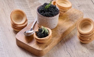 Siyah havyar, küçük bir seramik kap ve küçük bir tart, tahta bir tahtanın üzerine yerleştirilmiş. Kompozisyonda bir kaşık dolusu boş tart ve biberiye filizi yer alıyor..