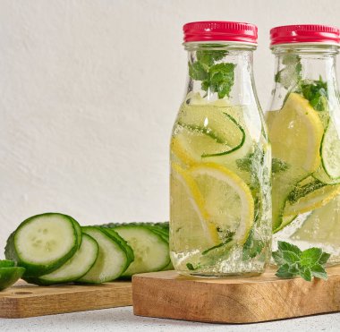 Kırmızı kapaklı cam şişeler, içinde salatalık, limon dilimleri ve nane yaprakları bulunan detoks suyla dolu beyaz masa.