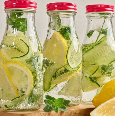 Kırmızı kapaklı cam şişeler, içinde salatalık, limon dilimleri ve nane yaprakları bulunan detoks suyla dolu beyaz masa.