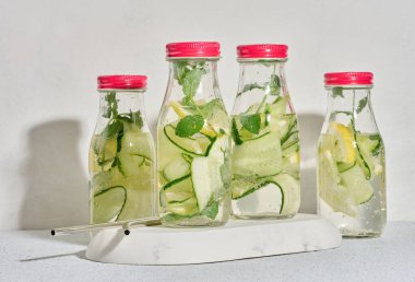 Kırmızı kapaklı cam şişeler, içinde salatalık, limon dilimleri ve nane yaprakları bulunan detoks suyla dolu beyaz masa.